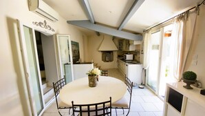 A dining area with a round table, chairs, a vase with flowers, a kitchen in the background, and a window with curtains. - Villa Provençale (La Cadière-d'Azur)
