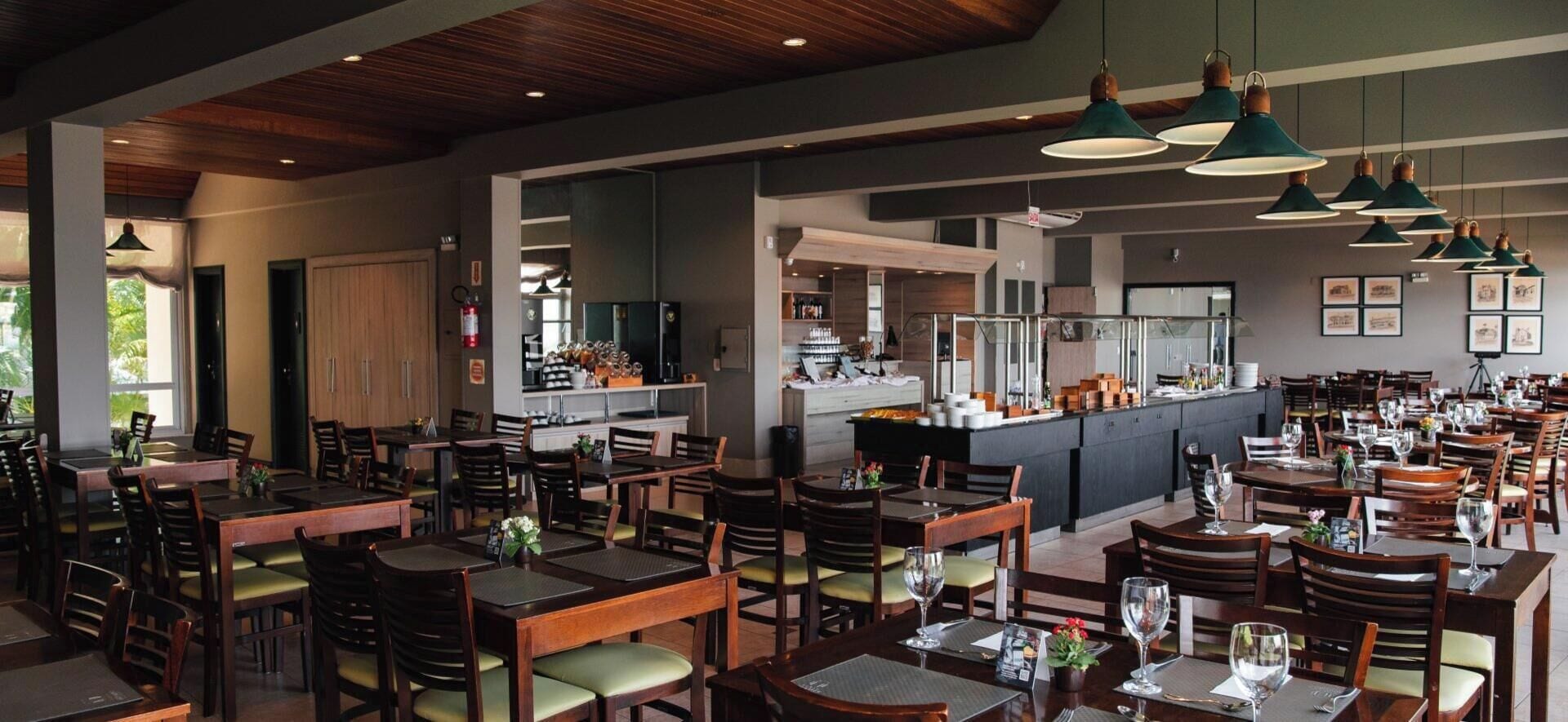 Un restaurant doté de plusieurs tables et chaises, d’un espace cuisine et d’un éclairage suspendu.