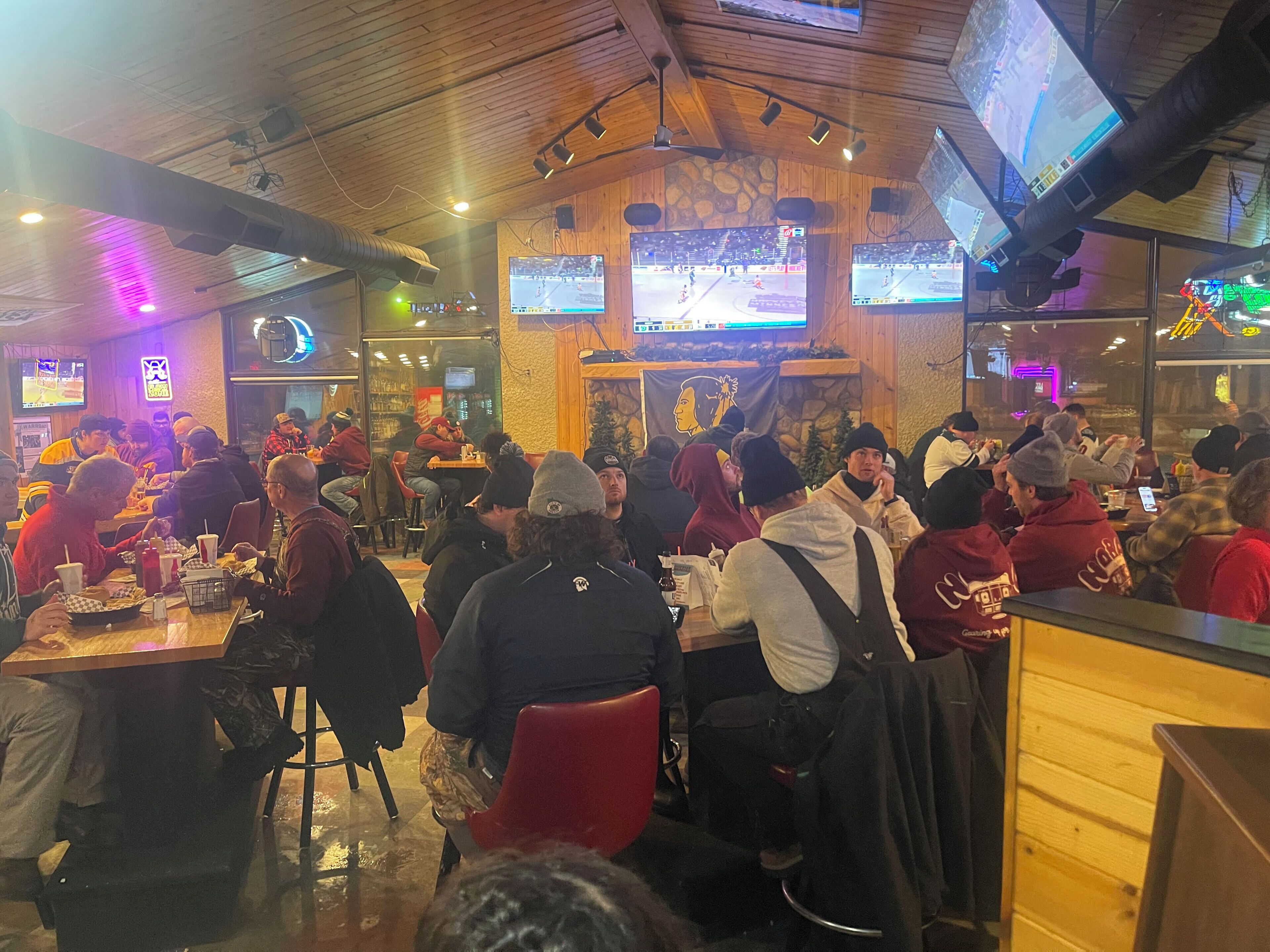A crowded indoor dining area with multiple tables and chairs, wooden walls, and ceiling-mounted lighting.