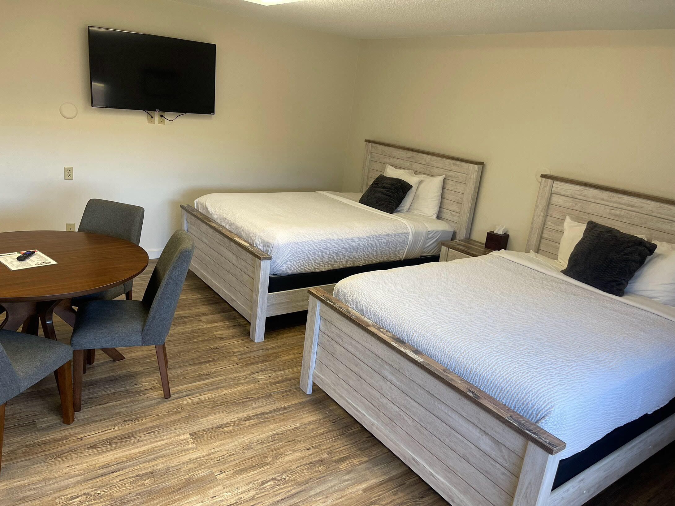 A hotel room with two single beds, a wooden dining table with chairs, and a flat-screen TV mounted on the wall.