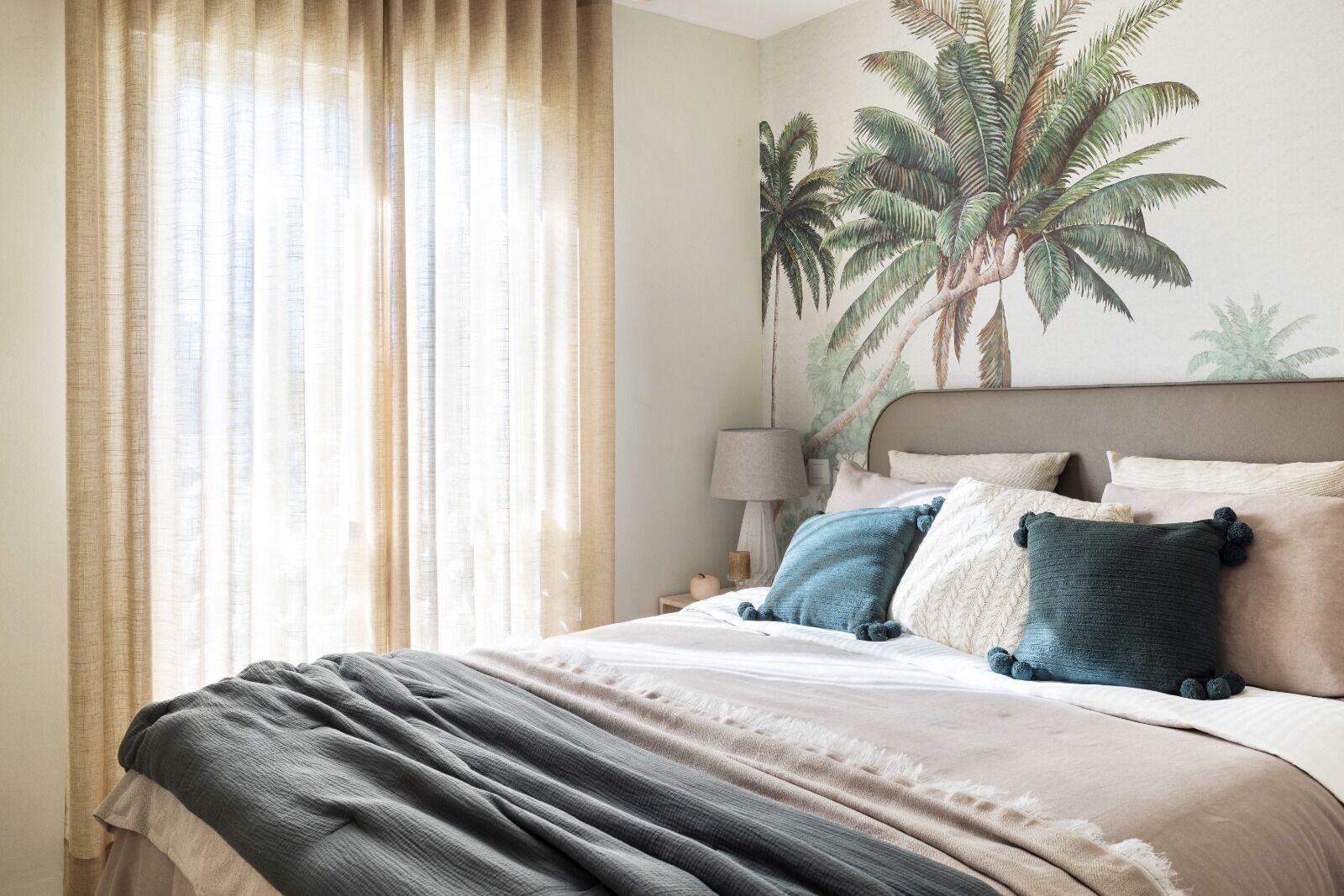 Een slaapkamer met een bed, nachtkastje, lamp en een muurschildering van palmbomen.