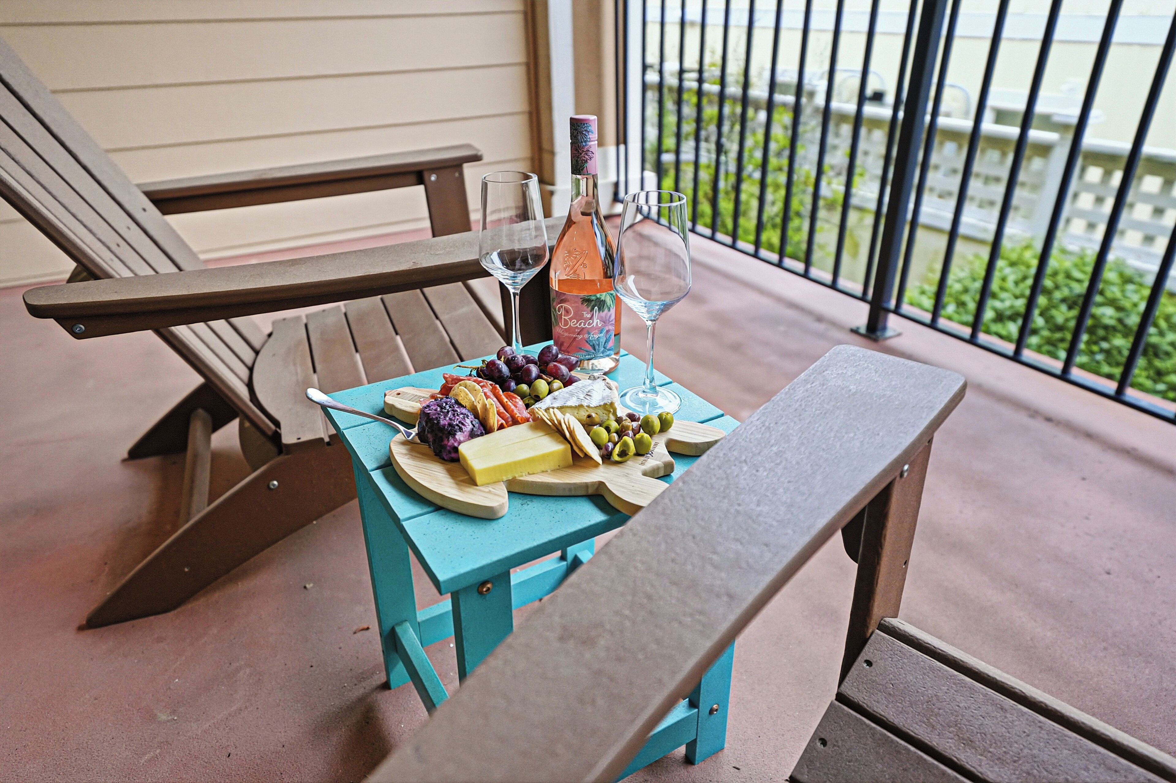 A wooden bench and table with a bottle of wine, glasses, and a variety of cheeses and olives on a balcony.