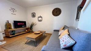 Ein modernes Wohnzimmer mit einer Couch, einem Holztisch, einem Fernseher mit der Anzeige 'NETFLIX', einer Uhr und einem Logo an der Wand.