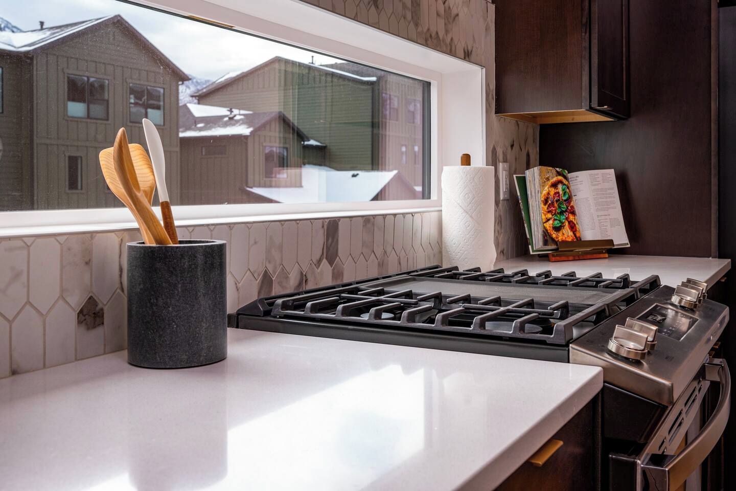 A modern kitchen with a gas stove, a window with a view of houses, and a paper towel holder.