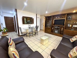 A living room with a brown leather sofa, a glass coffee table, a dining table with chairs, a TV on a wooden stand, and a potted plant. - Aparts4Days Miraflores Down Town 1 or 3 Bedroom Apartment (Lima)