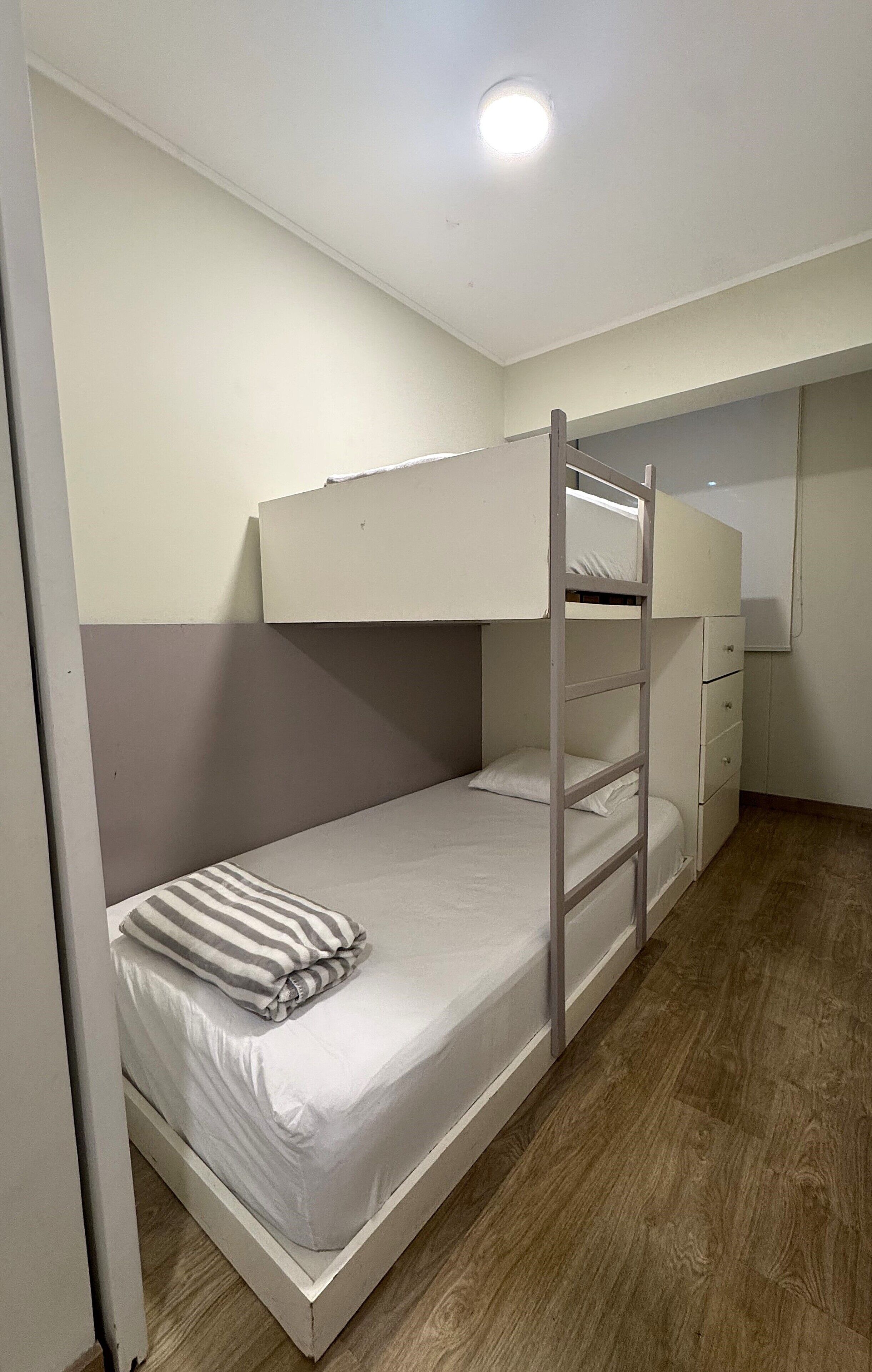 Un llit superposat amb escala, un taulell petit i un moble amb calaixos.