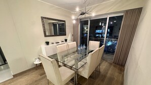 A modern dining area with a glass table and white chairs, a large mirror, and a view of the city at night. - Aparts4Days Miraflores Down Town 1 or 3 Bedroom Apartment (Lima)
