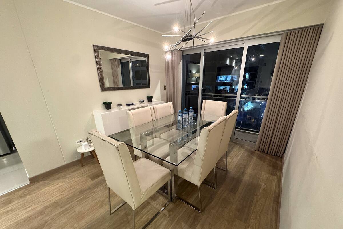 Un espace de repas moderne avec une table en verre et des chaises blanches, un grand miroir et une vue sur la ville la nuit.