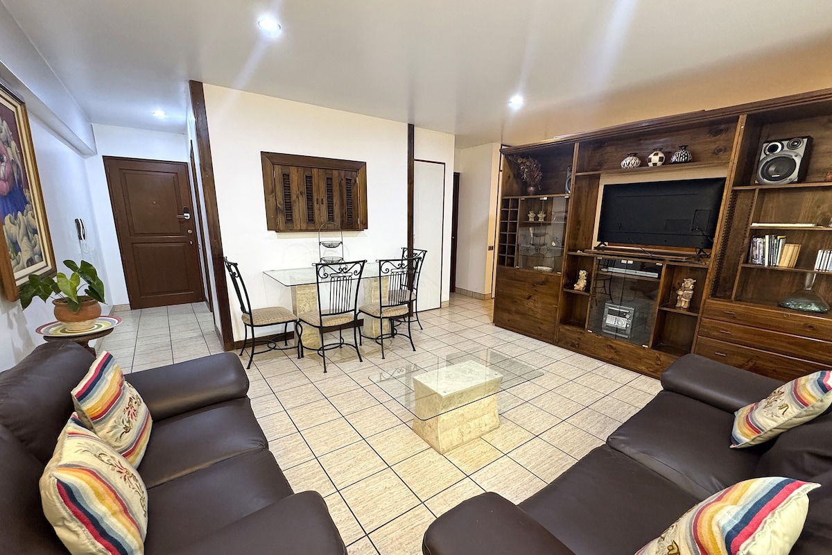 A living room with a sofa set, a dining table with chairs, a TV stand with shelves, and a door.