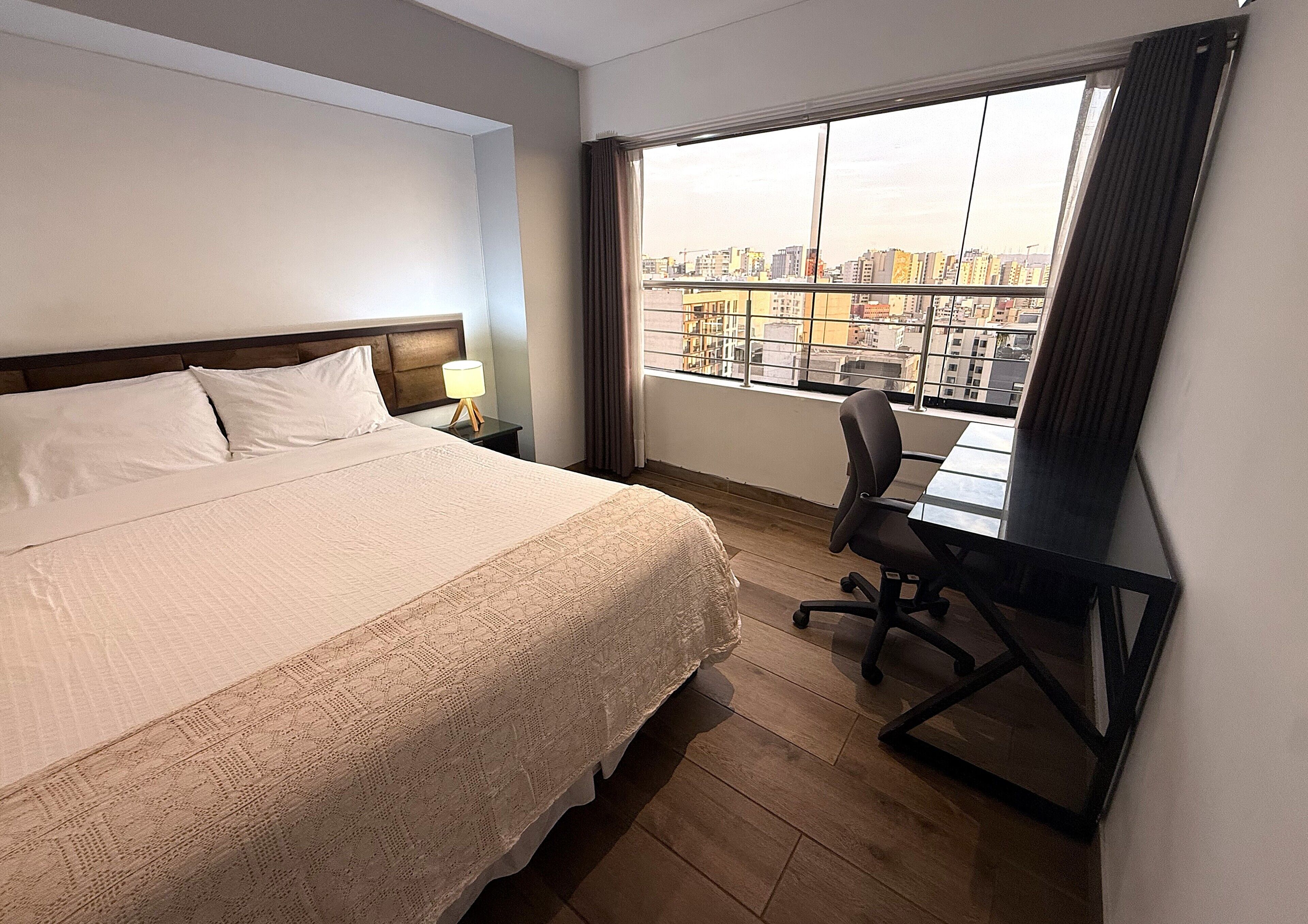 A hotel room with a bed, a desk, a chair, a nightstand, and a large window with a city view.