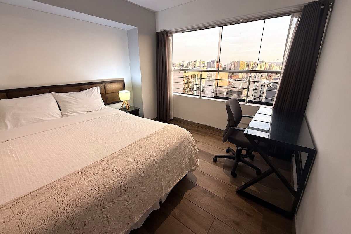 Habitación de hotel con cama, escritorio, silla, mesita de noche y un amplio ventanal con vistas a la ciudad.