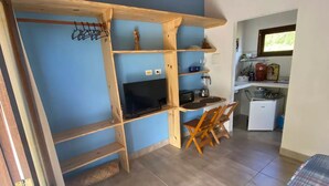 Una habitación pequeña con escritorio de madera, una silla, televisor y zona de kitchenette.