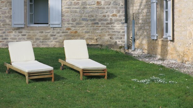 Dos sillones de madera con cojines blancos sobre un césped frente a un edificio de piedra con ventanas y contraventanas.