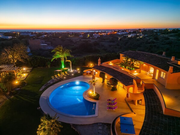 A large swimming pool with a sunken seating area, surrounded by a well-lit patio with outdoor furniture.