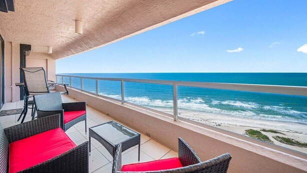 Ein Balkon mit Korbmöbeln und roten Kissen mit Blick auf Strand und Meer.