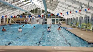 An indoor swimming pool with multiple lanes, a roof structure, and a poolside area with chairs and a lifeguard stand.