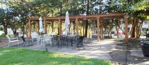 Relax in a serene outdoor setting surrounded by trees, with a pergola providing shade and comfortable seating for dining or socializing.
