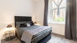 Un dormitorio con cabecera capitonada, colcha estampada, lámpara de noche y ventana con cortinas.