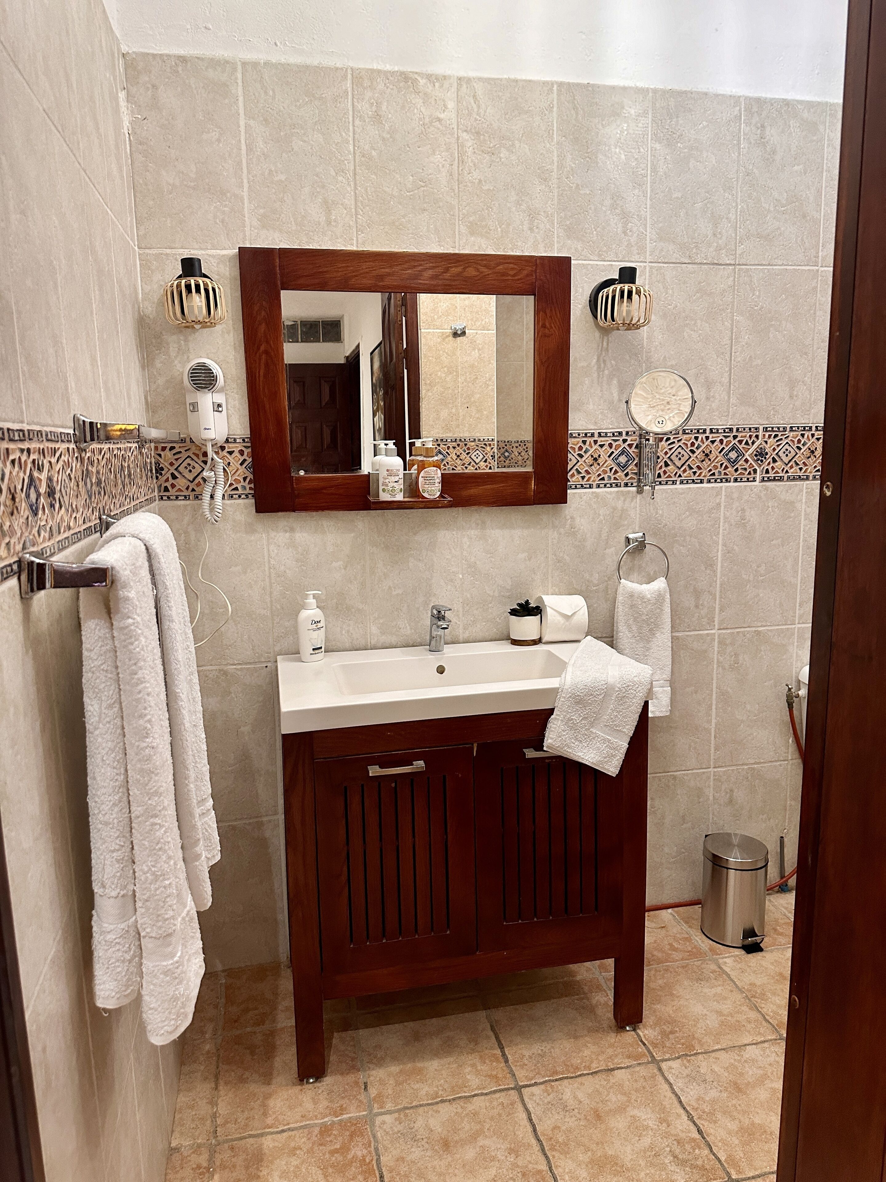 A bathroom with a wooden vanity, a white sink, and a mirror above it. There are towels hanging on the wall and a trash can in the corner.