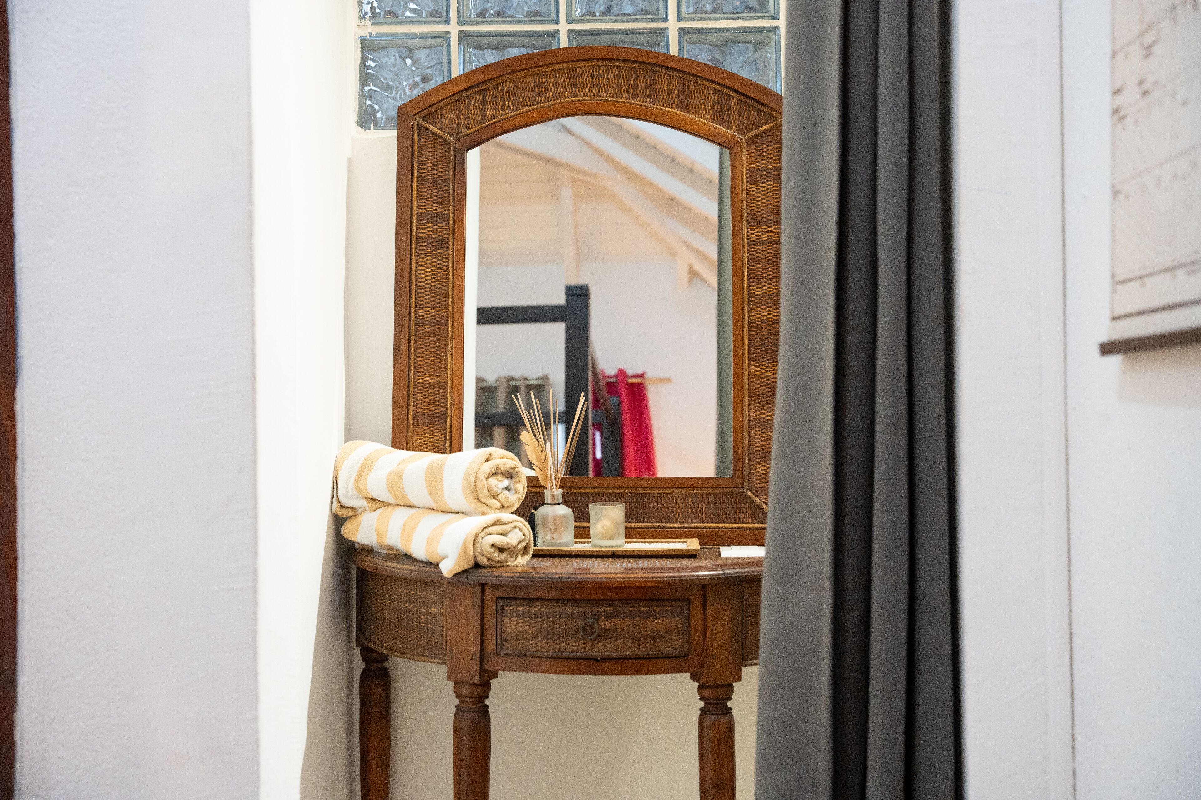 A wooden vanity with a mirror, a rolled towel, and a small vase with reeds.