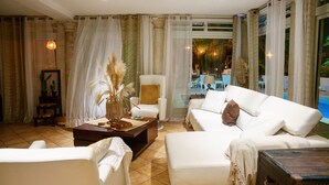 A spacious living room with a white sofa, a coffee table, and a chair. There is a large window with curtains, a view of the pool outside, and a framed picture on the wall. - Villa Kapresse du 978 (Marigot)