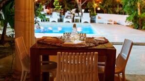 An outdoor dining area with a table set for two, surrounded by white chairs, near a pool and palm trees. - Villa Kapresse du 978 (Marigot)