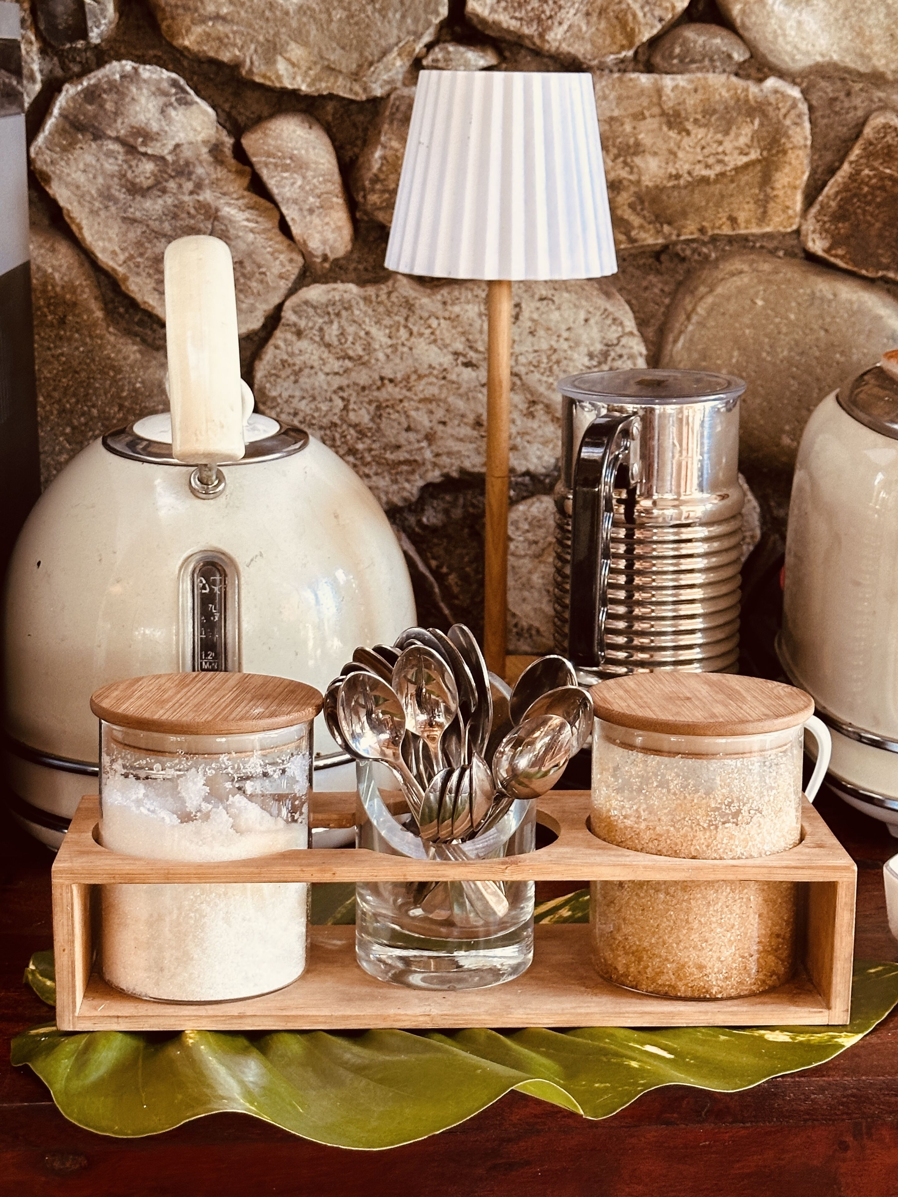 A kitchen counter with a stone wall background, a white kettle, a silver pitcher, and two wooden containers with lids.