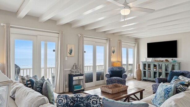 A spacious living room with a large window overlooking the ocean, a ceiling fan, and a television mounted on the wall.