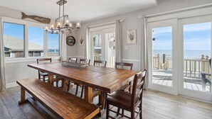 A spacious dining area with a large wooden table, benches, and chairs. There is a chandelier above the table and a view of the ocean through large windows.