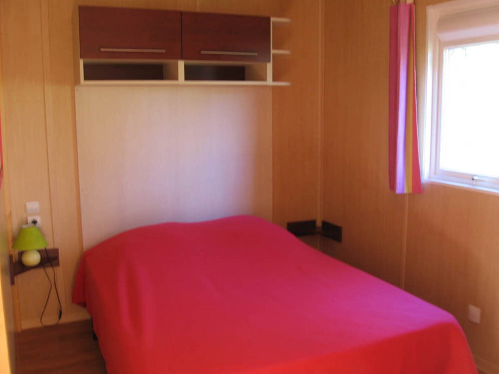 A bedroom with a bed, wooden walls, a window with pink curtains, and a green lamp on a shelf.