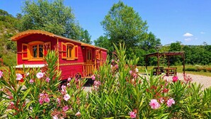A red wooden house with large windows and a wooden porch, surrounded by pink flowers and greenery. - 4/5 people trailers (MONTREAL)