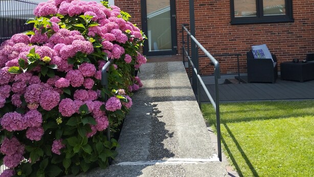 Una casa in mattoni con portico coperto, un'apertura di garage ben visibile e un'alta siepe di fiori rosa.