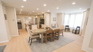 Un vaste espace dédié à la salle à manger, avec une grande table et des chaises en bois, une cuisine à l’arrière-plan et une vue sur l’extérieur grâce à de grandes fenêtres.
