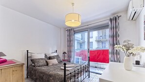 Un dormitorio con cama, mesita de noche, lámpara, ventana con cortinas y vistas a edificios.