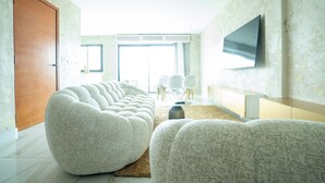 A modern living room with a large sectional sofa, a flat-screen TV mounted on the wall, and a large window providing natural light. - The Six Apparthotel (Abidjan)