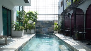 A swimming pool with a stone wall and a waterfall feature, surrounded by potted plants and outdoor seating. - The Six Apparthotel (Abidjan)