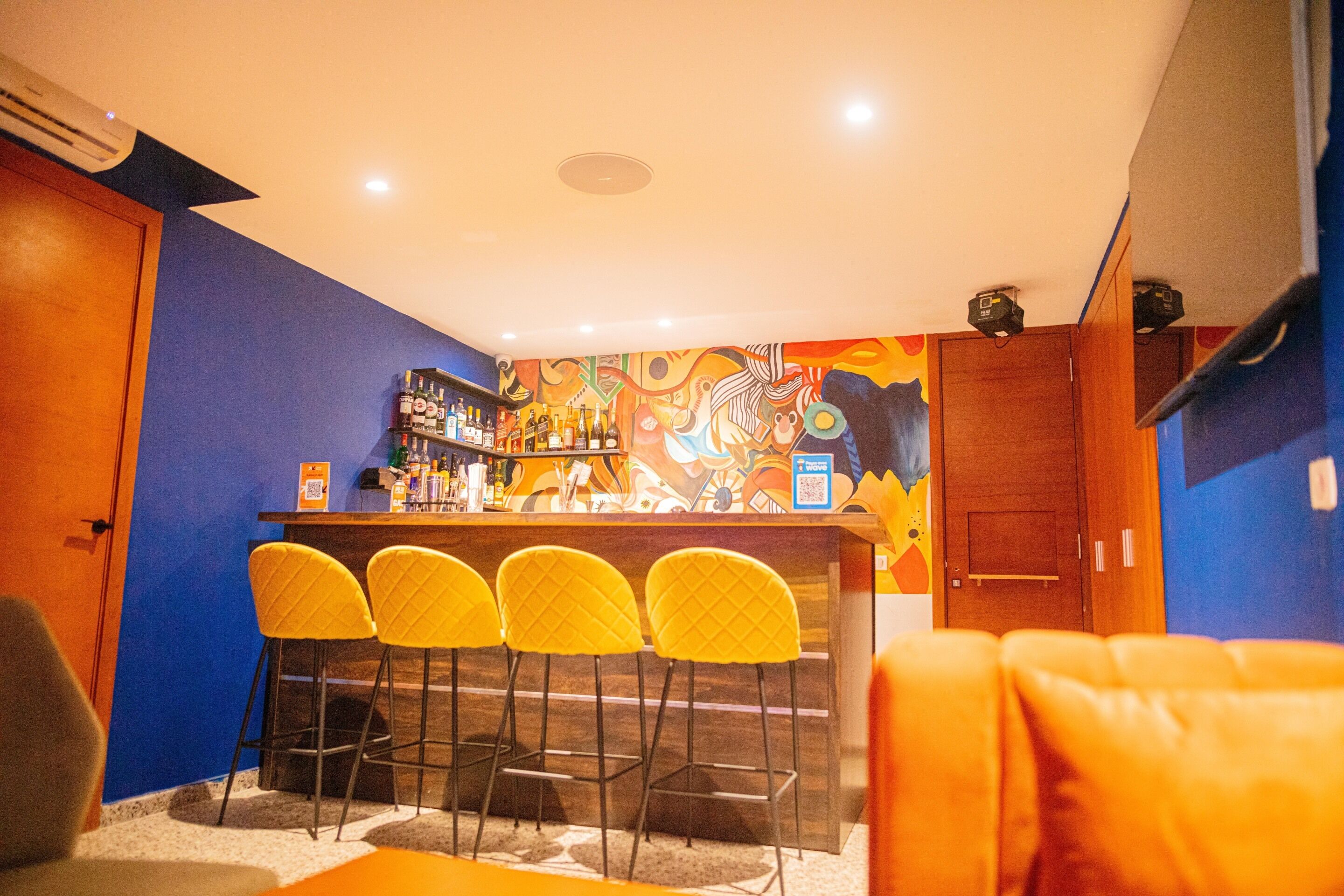 Un espace bar avec un comptoir, quatre chaises jaunes et une fresque colorée sur le mur.