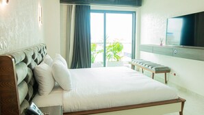 A hotel room with a bed, a TV, a bench, and a view of greenery outside. - The Six Apparthotel (Abidjan)