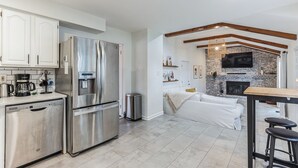 Una cocina moderna con electrodomésticos de acero inoxidable, lavavajillas y cafetera. En la sala de estar, hay un sofá blanco y una chimenea.