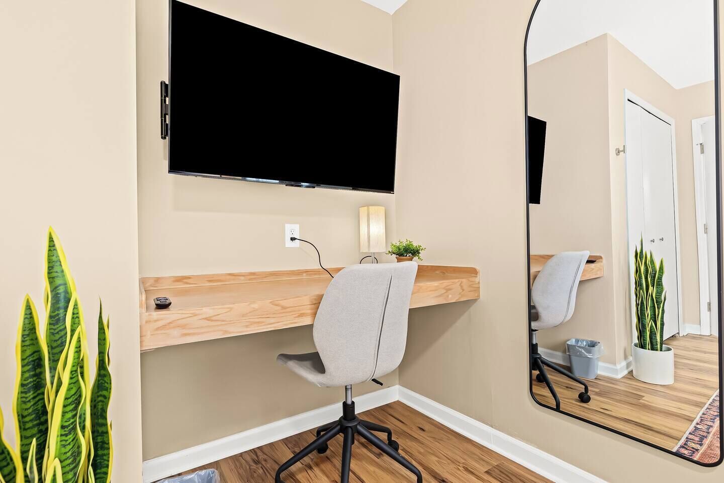 Ein modernes Zimmer mit einem wandmontierten Fernseher, einem Holztisch, einem grauen Bürostuhl, einem kleinen Topfpflanzen und einem großen Spiegel, der das Zimmer reflektiert.