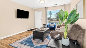 A living room with a flat-screen TV, a coffee table, a blue armchair, a brown sofa, a large green plant, and a patterned rug. - Brand New! Lower Flat 1BR Near Downtown, Roseville (Roseville)