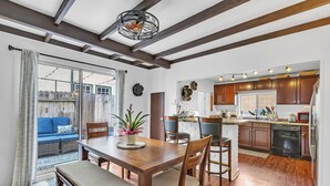 A modern kitchen with wooden cabinets, a dining area with a wooden table and chairs, and a view of a blue outdoor seating area. - Historic "Spanish Jewel" at HOLLYWOOD BEACH (Oxnard)