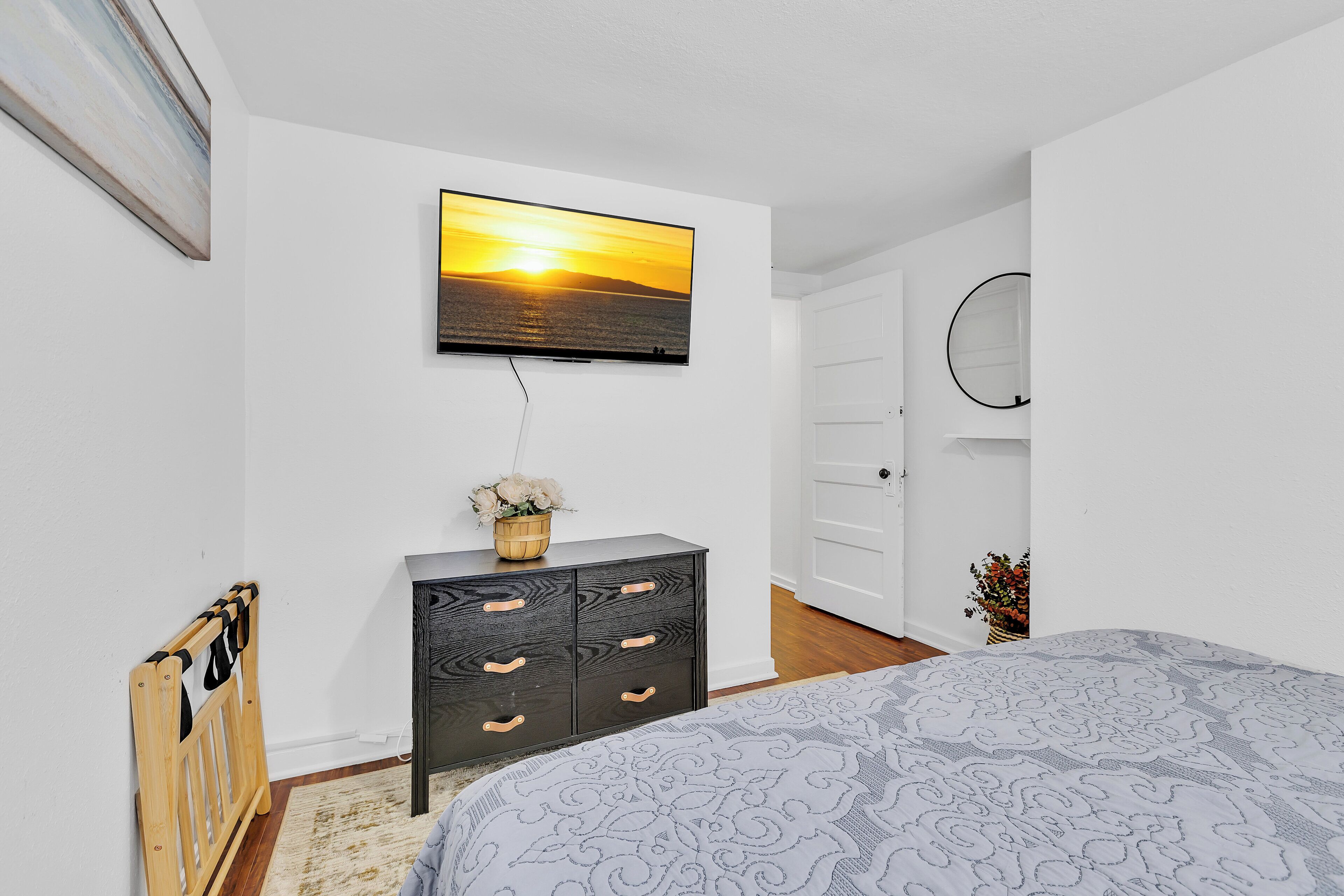 A bedroom with a bed, a dresser, a TV mounted on the wall, and a door leading to another room.