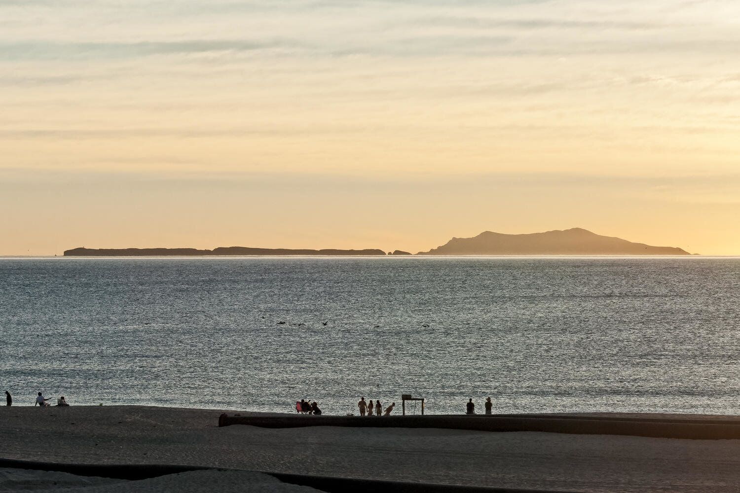Een strand met een rustige zee, een eiland in de verte en mensen die langs de kust wandelen.