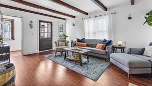 A living room with a sofa, armchairs, a coffee table, and a rug. There is a door, a window with curtains, and a plant. - Historic "Spanish Jewel" at HOLLYWOOD BEACH (Oxnard)
