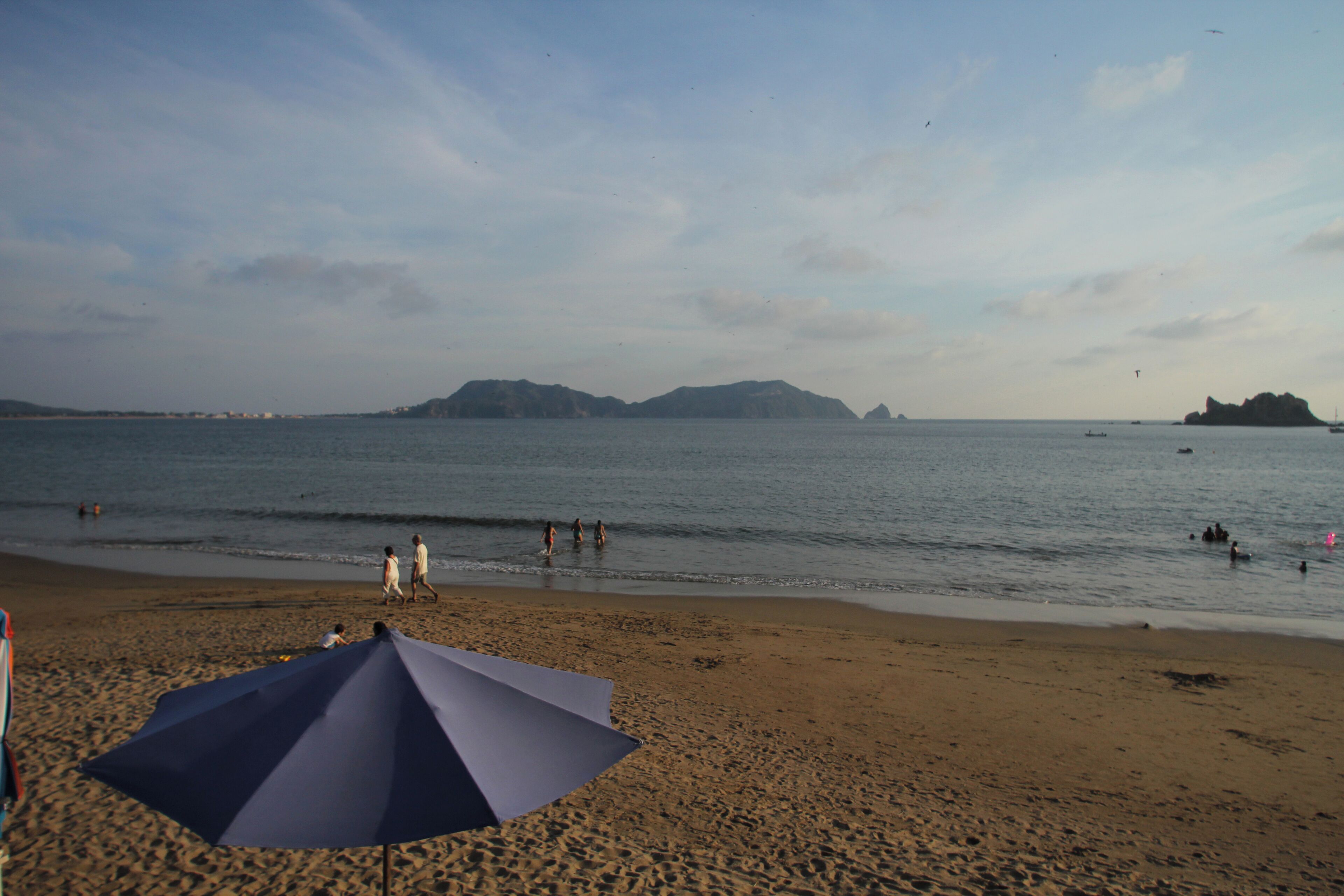 A beach with a large umbrella, people walking, and boats in the water.