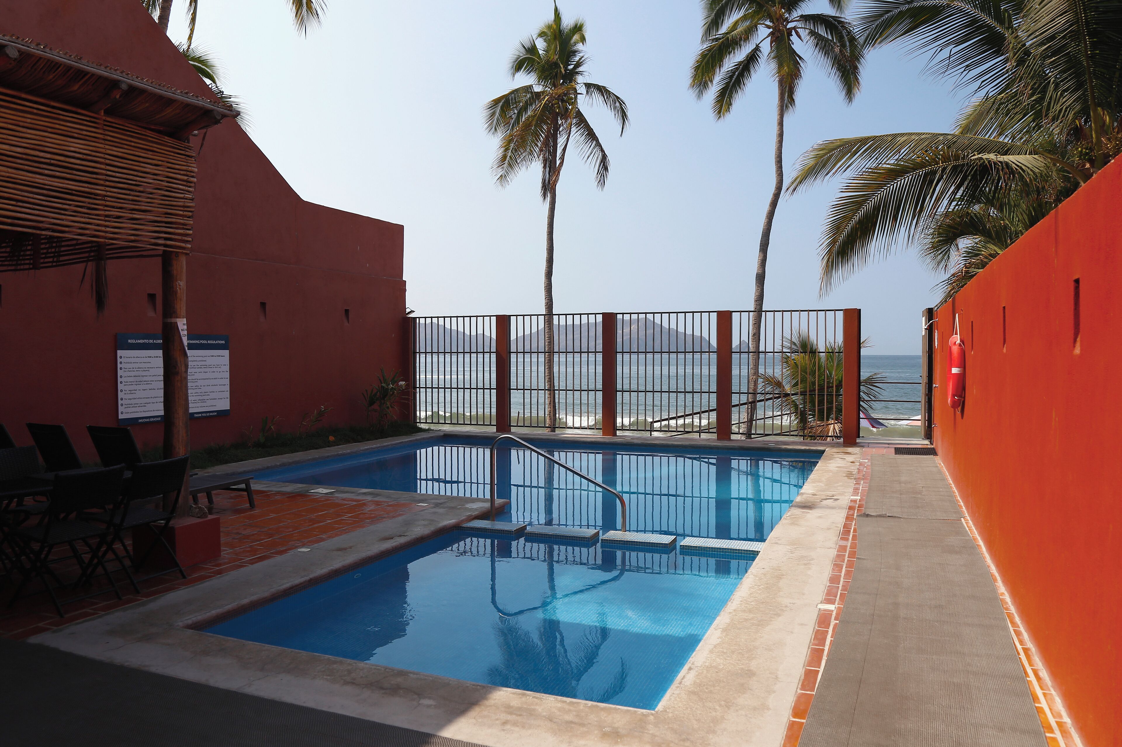 Zona de piscina con vista al océano, palmeras y un muro rojo.