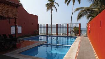 Zona de piscina con vista al océano, palmeras y un muro rojo.