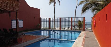 A pool area with a view of the ocean, palm trees, and a red wall.