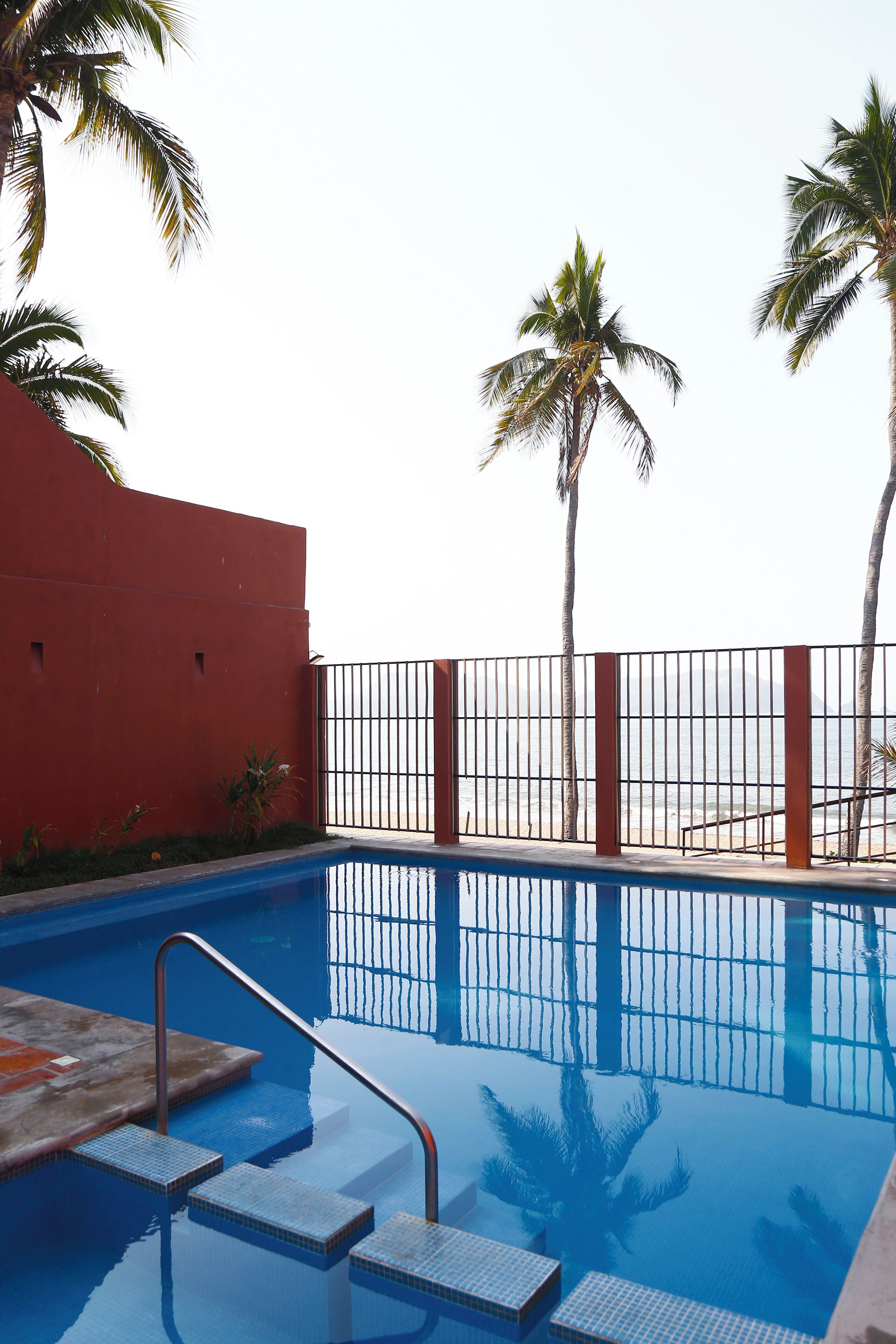 An outdoor swimming pool with steps leading into the water, surrounded by a red wall and a metal fence.
